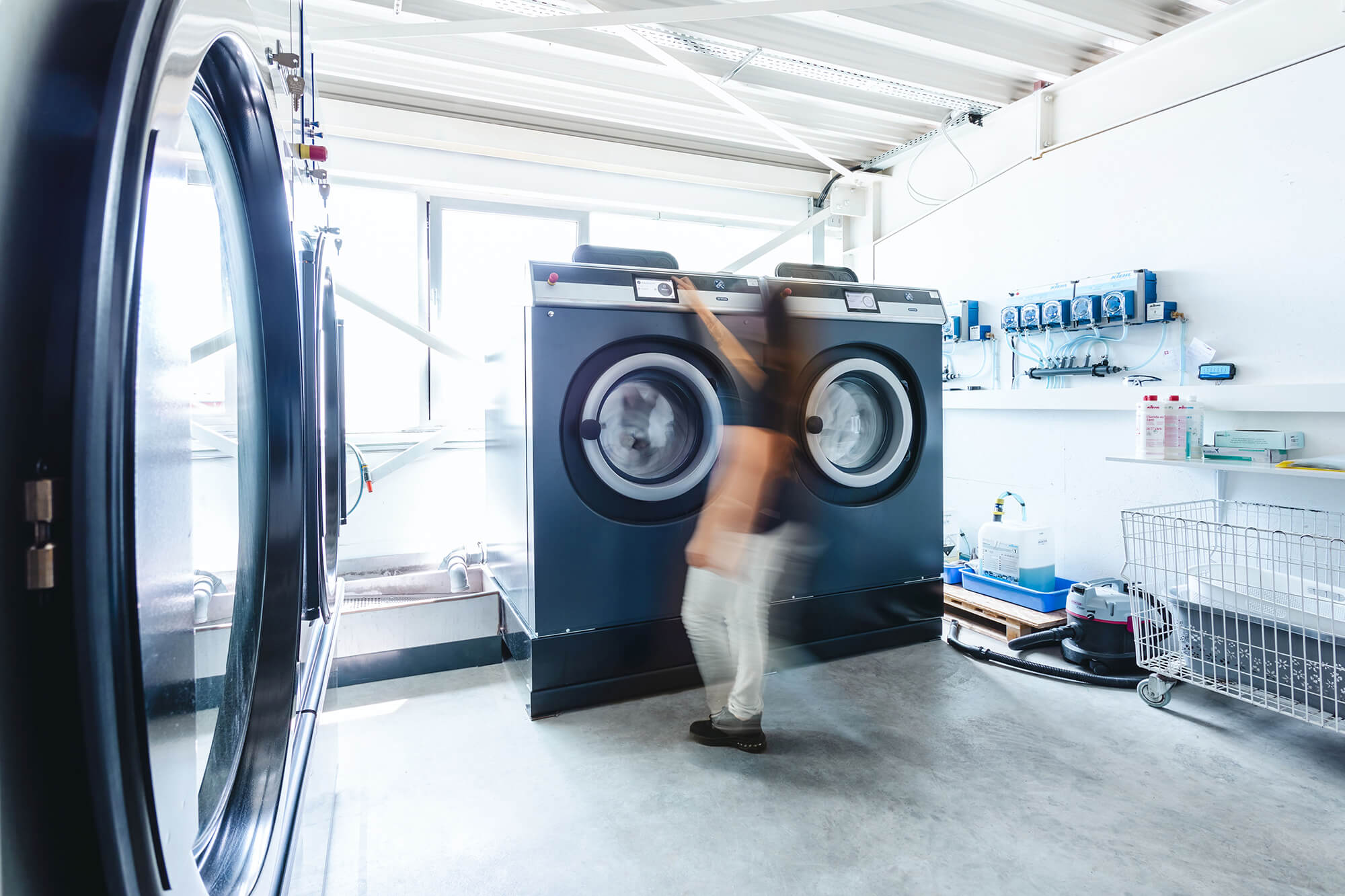Person bedient industrielle Waschmaschine in einem modernen, hellen Waschraum mit technischer Ausstattung.