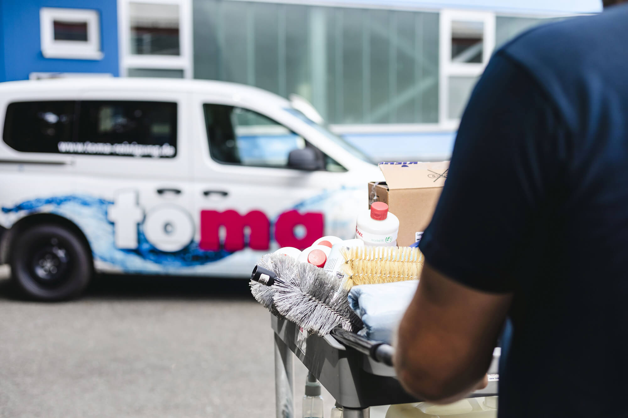 Reinigungskraft mit Reinigungswagen vor einem Firmenfahrzeug der Toma Gebäudereinigung – ausgestattet mit Putzmitteln, Bürsten und Reinigungszubehör.